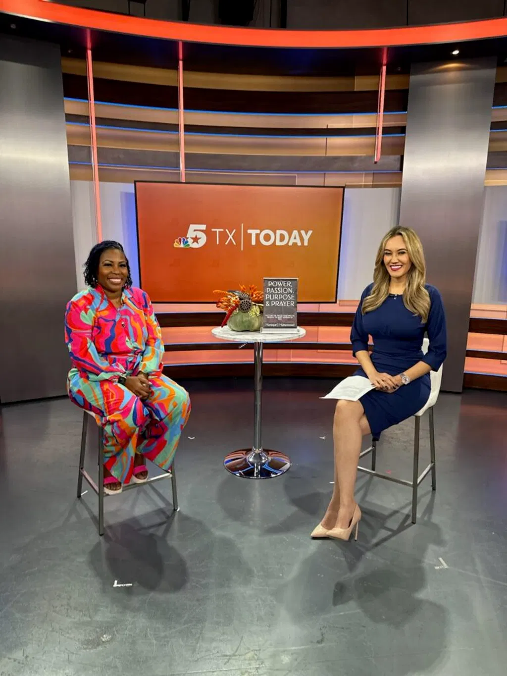 Two women sit on tall stools in a TV studio set with an orange TX Today backdrop, a small table between them holding a book and decorative item.